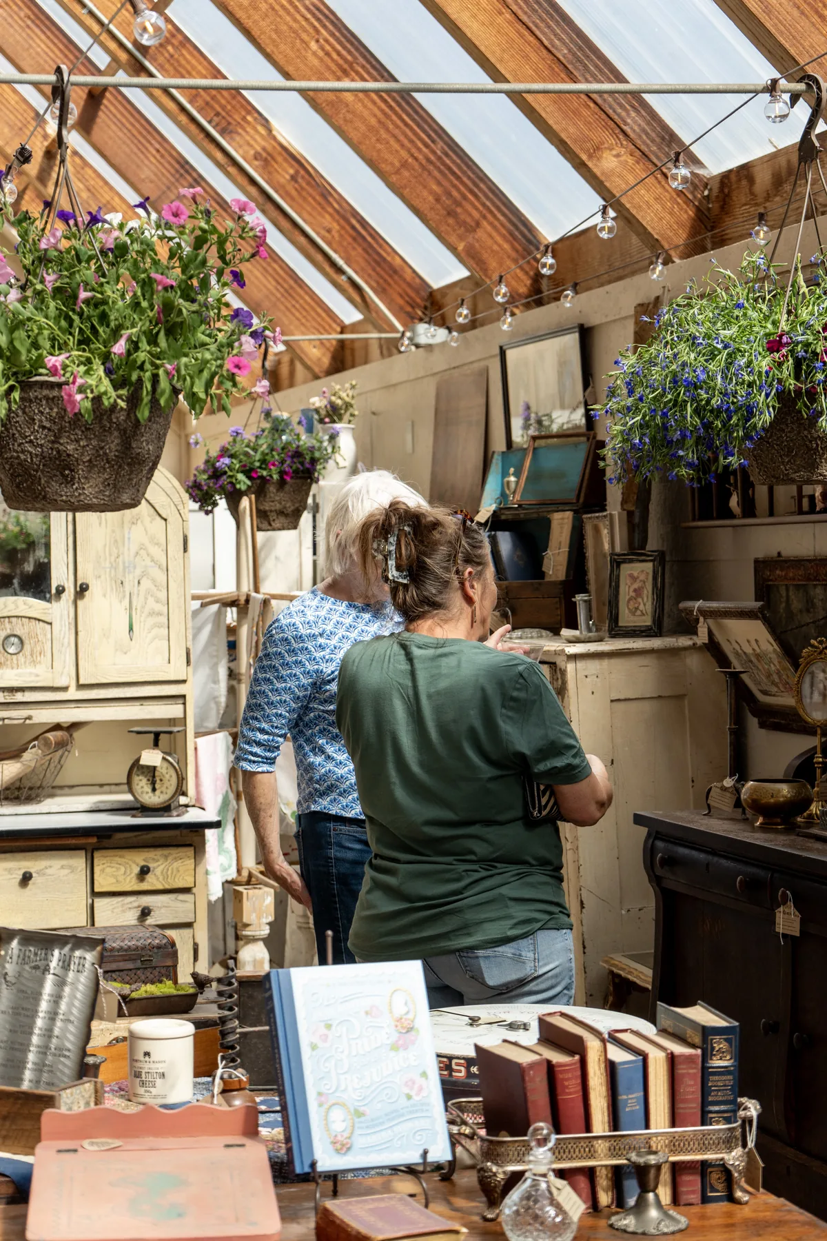 Friends browsing collected antiques beneath hanging flower baskets during a greenhouse gathering