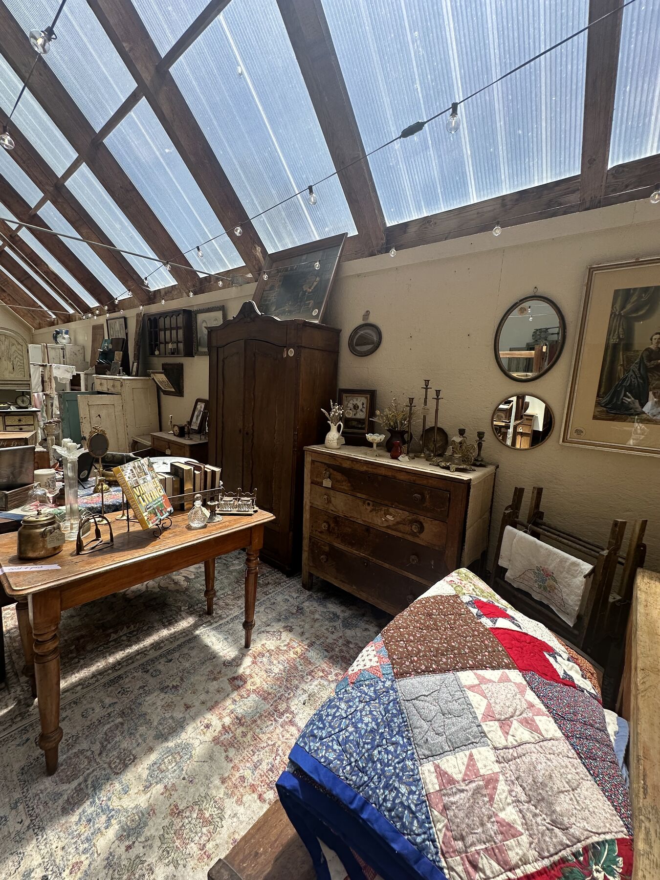 A quiet greenhouse corner arranged with antique furniture, mirrors, and folded quilts